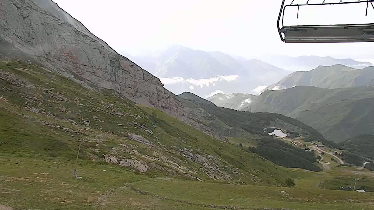 A mountainside with sparse vegetation and rocky outcrops overlooks a valley with a small lake and distant mountains under a mostly cloudy sky.