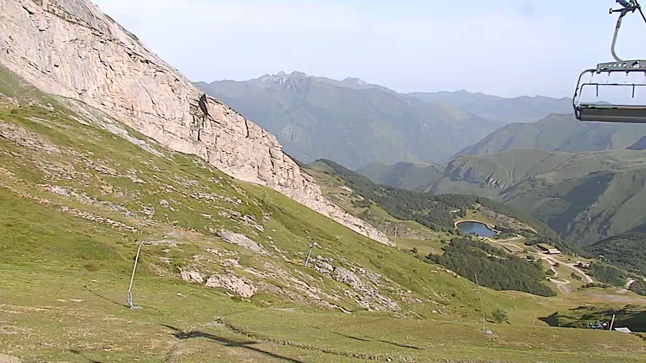 A grassy mountain slope with a rocky cliff descends toward a valley containing a small lake and a ski lift chair is visible in the upper right corner under a clear sky.