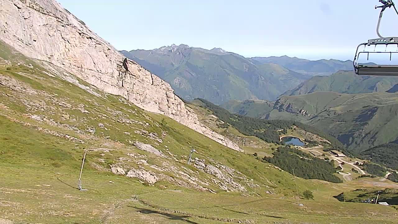 A chairlift ascends a grassy mountain slope overlooking a valley with distant mountains under a clear sky.