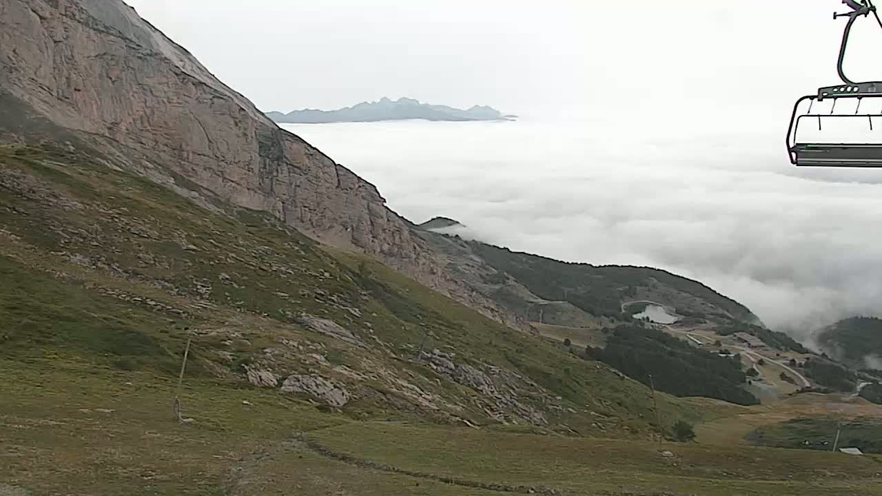 A mountainside with sparse vegetation and a rocky cliff overlooks a valley shrouded in fog, with a chairlift in the upper right corner.