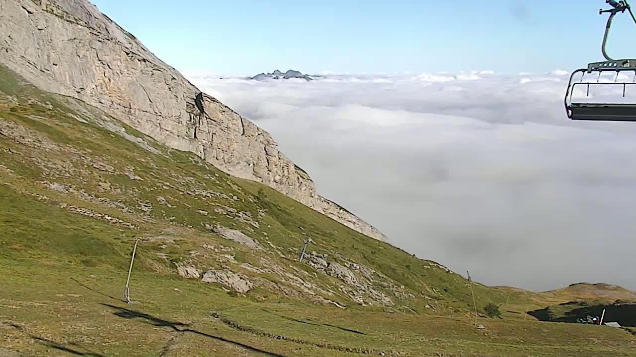 A grassy mountainside with a rocky cliff overlooking a valley filled with low clouds, a chairlift is visible in the upper right corner.