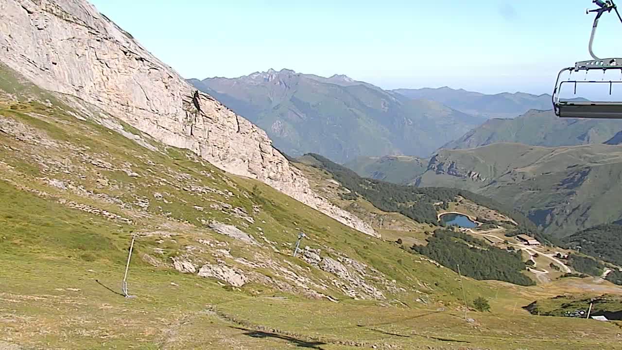 A chairlift ascends a grassy mountain slope overlooking a valley with a small lake, under a clear sky.