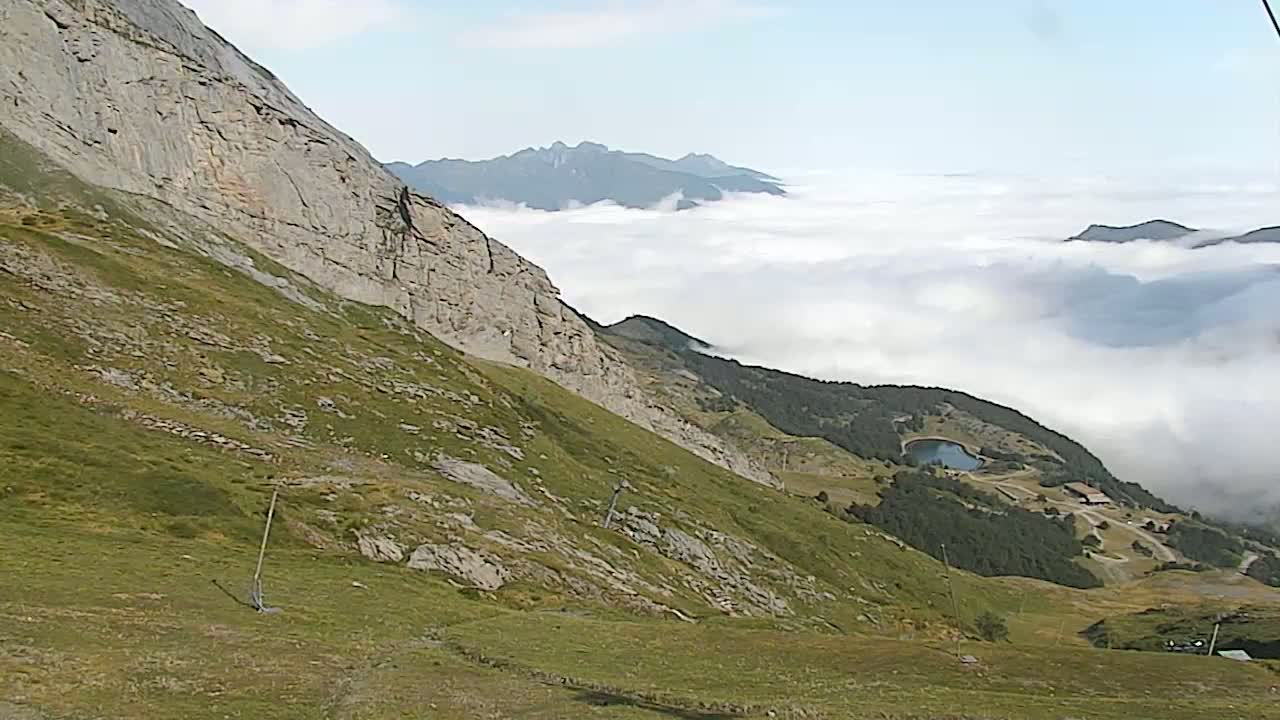 A mountainous landscape with grassy slopes and a rocky cliff overlooks a valley filled with low clouds and a small lake.