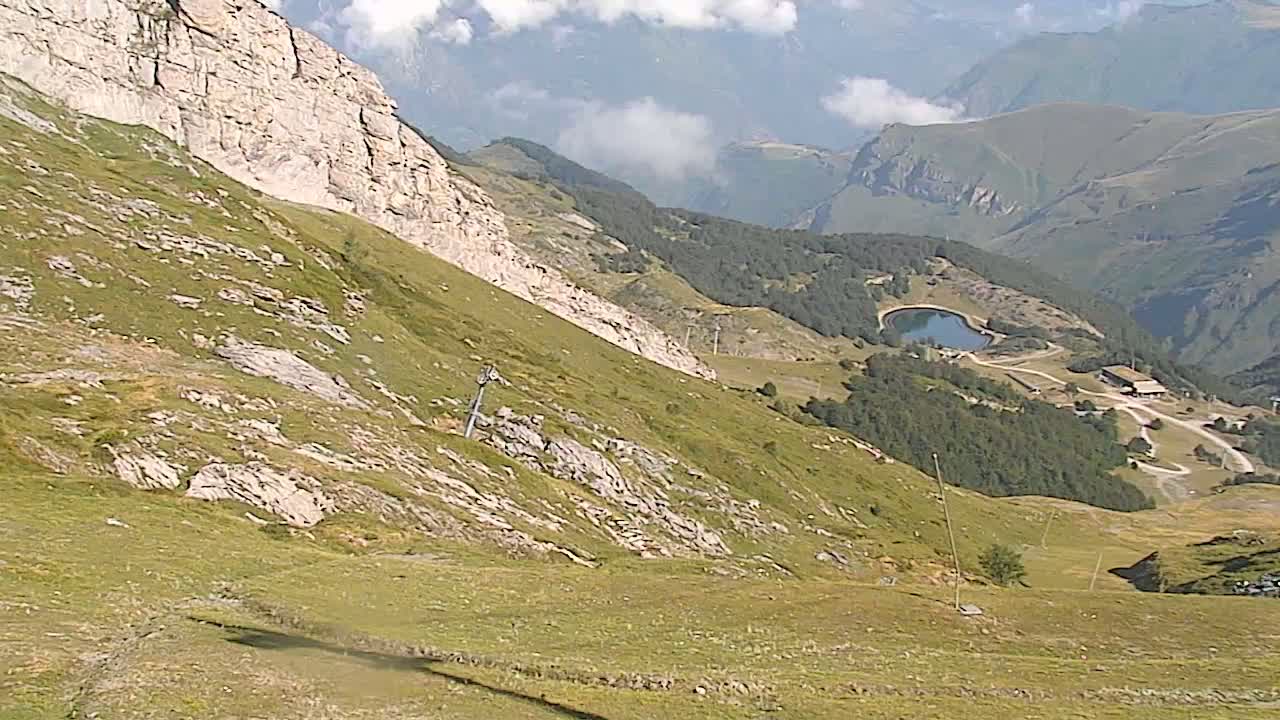 A grassy mountain slope descends toward a valley with a small lake and buildings, under partly cloudy skies.