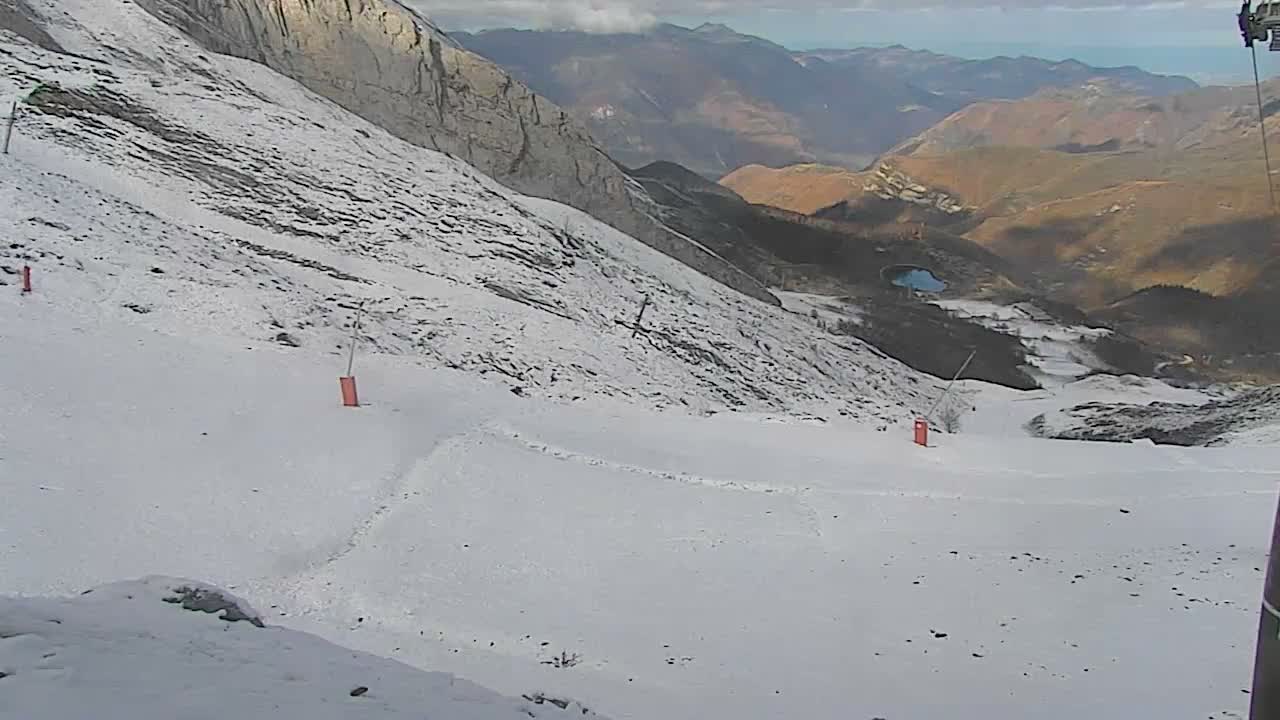 A snow-covered ski slope with orange markers descends towards a valley featuring a small blue lake and distant mountains under a partly cloudy sky.