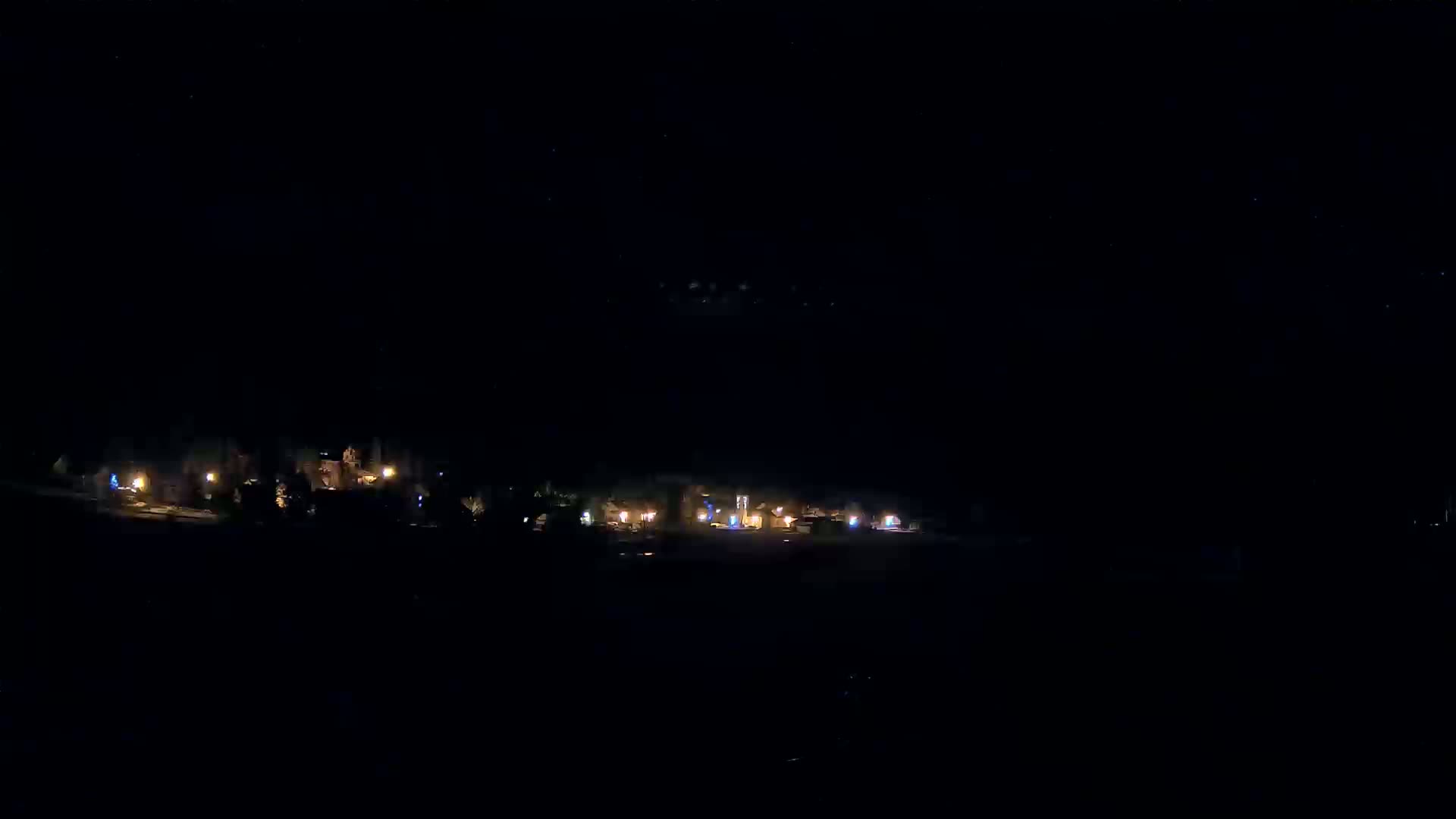 A nighttime long shot shows a sparsely lit cluster of buildings under a dark, clear sky.