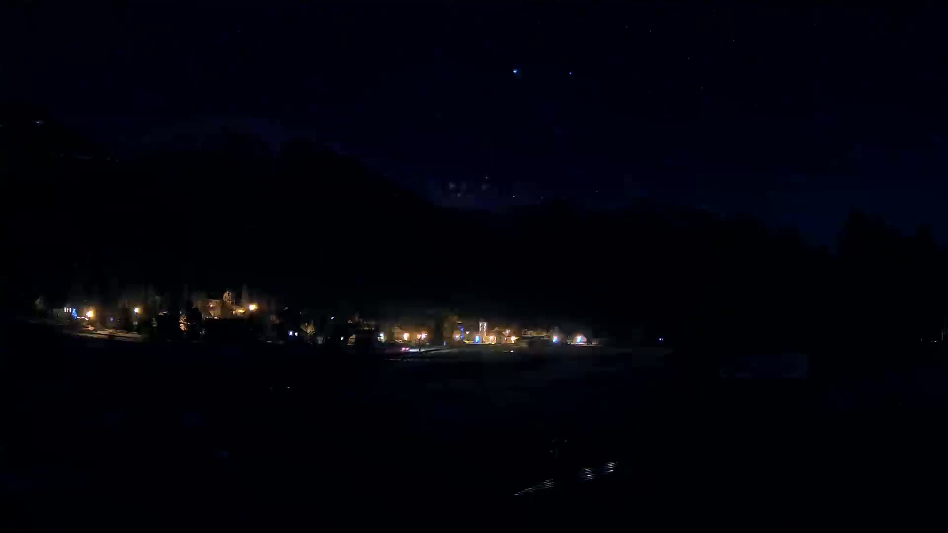 A small cluster of dimly lit buildings sits at the base of a dark mountain under a clear, starlit night sky.
