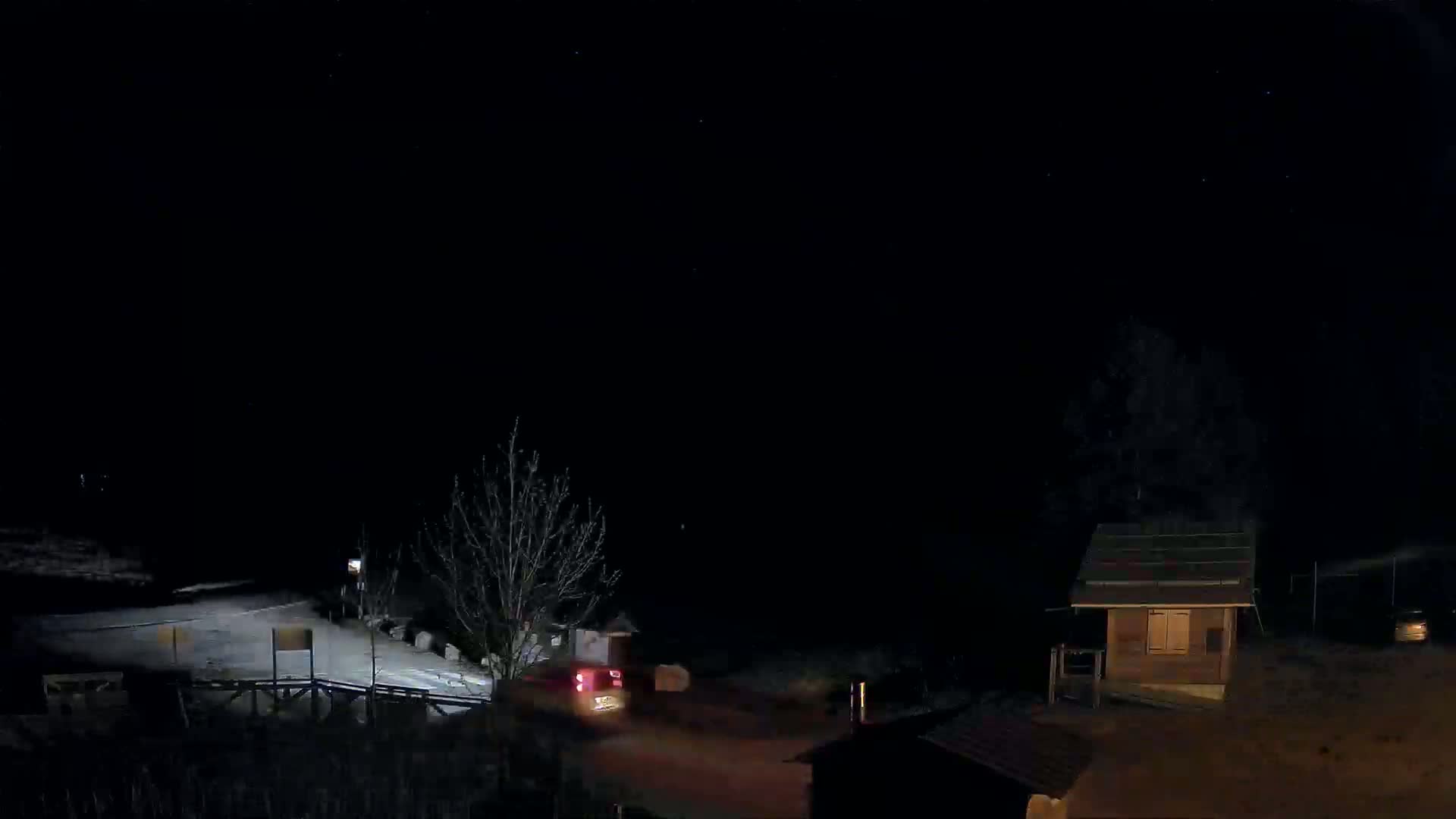 A nighttime scene shows a vehicle on a road near a small building and bare trees under a clear, dark sky.