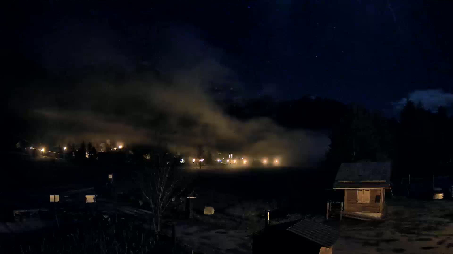 A nighttime scene shows a small, dimly lit town partially obscured by fog or low clouds under a dark, starry sky, with a small wooden building in the foreground.