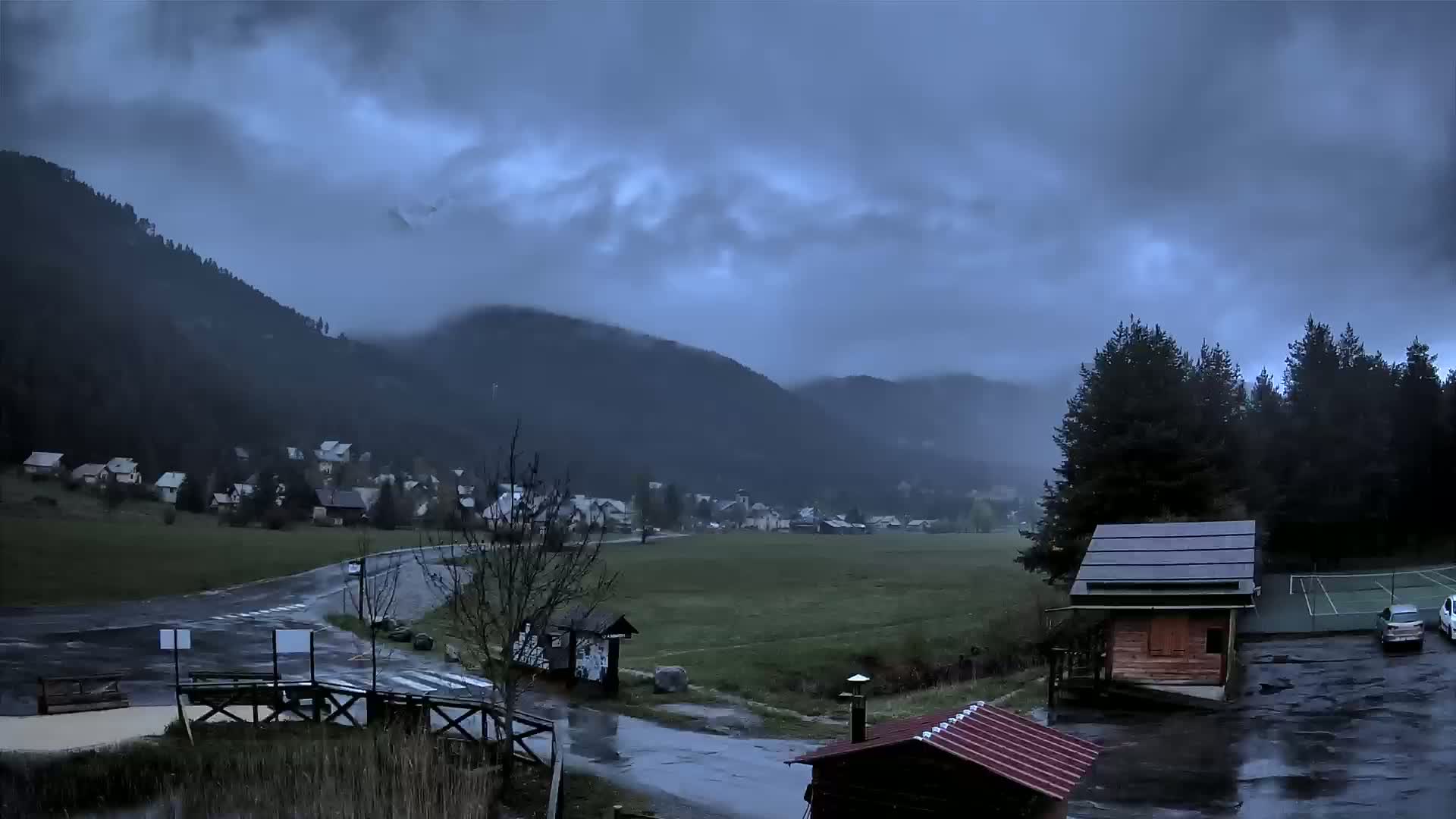 A small village nestled in a valley between dark, rain-covered mountains under a stormy sky.