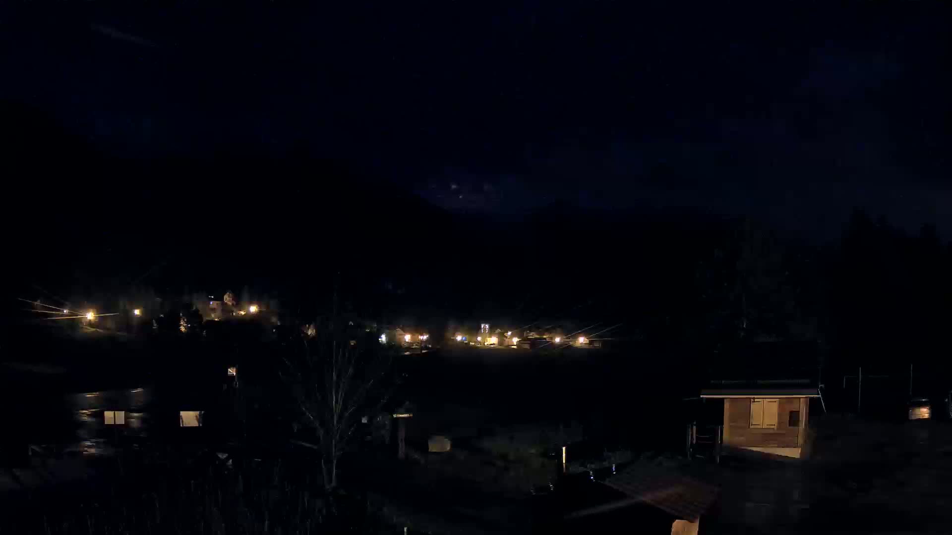 A nighttime view shows a small village nestled in a dark valley under a clear, starlit sky.