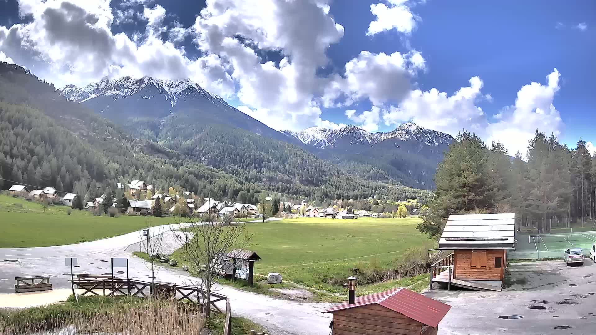 A small village nestled in a valley, surrounded by snow-capped mountains under a partly cloudy sky.