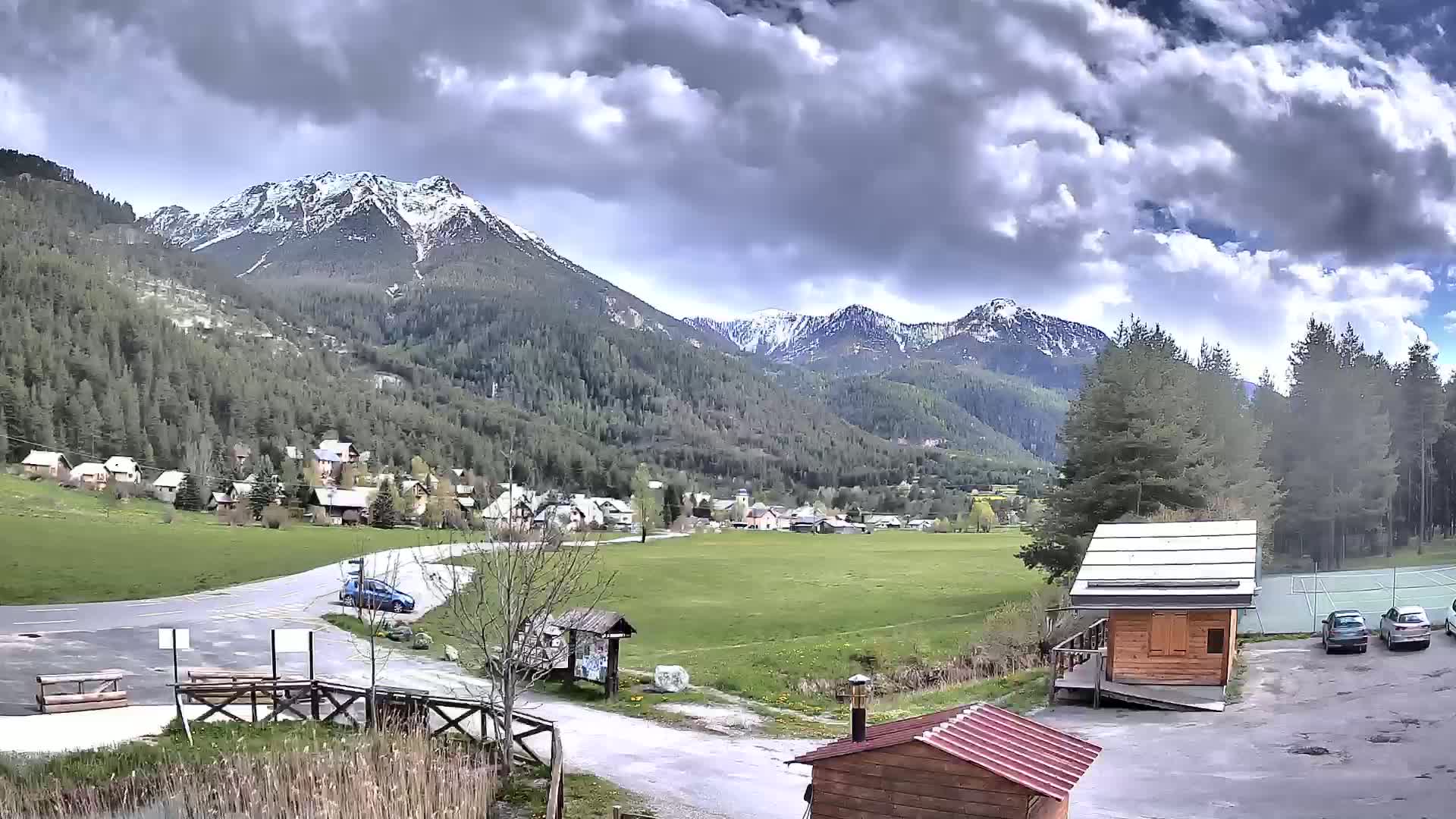 A partly cloudy day reveals a small village nestled in a valley between snow-capped mountains, with a few cars parked near a small wooden building.