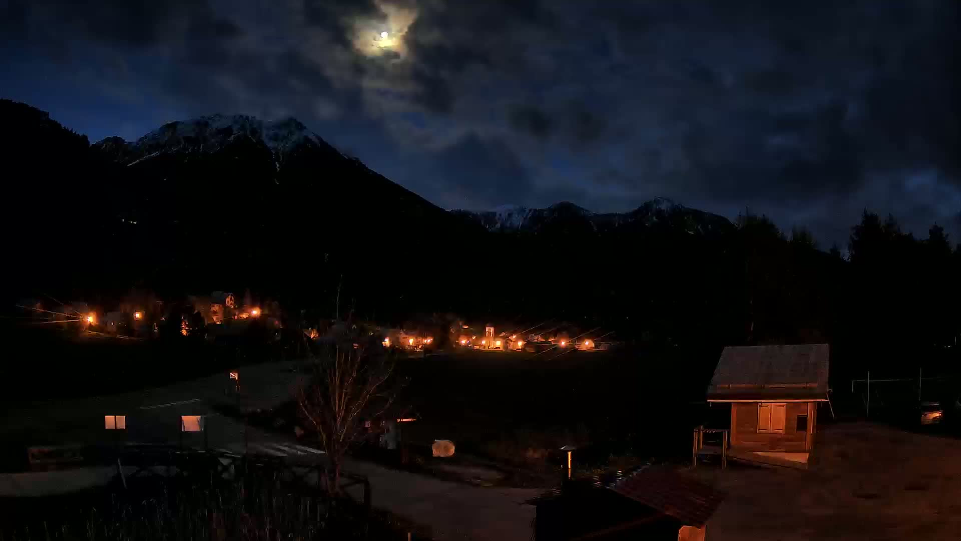 A moonlit night scene shows a small village nestled in a valley between snow-capped mountains.
