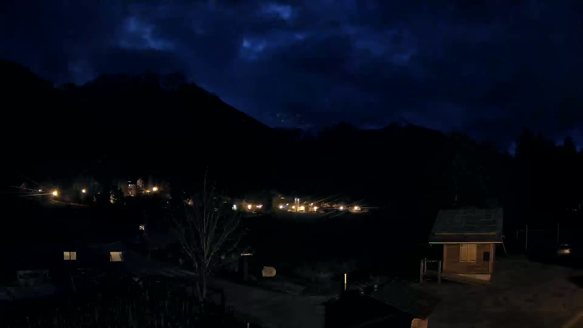 A nighttime view of a small village nestled in a dark valley under a partly cloudy sky.