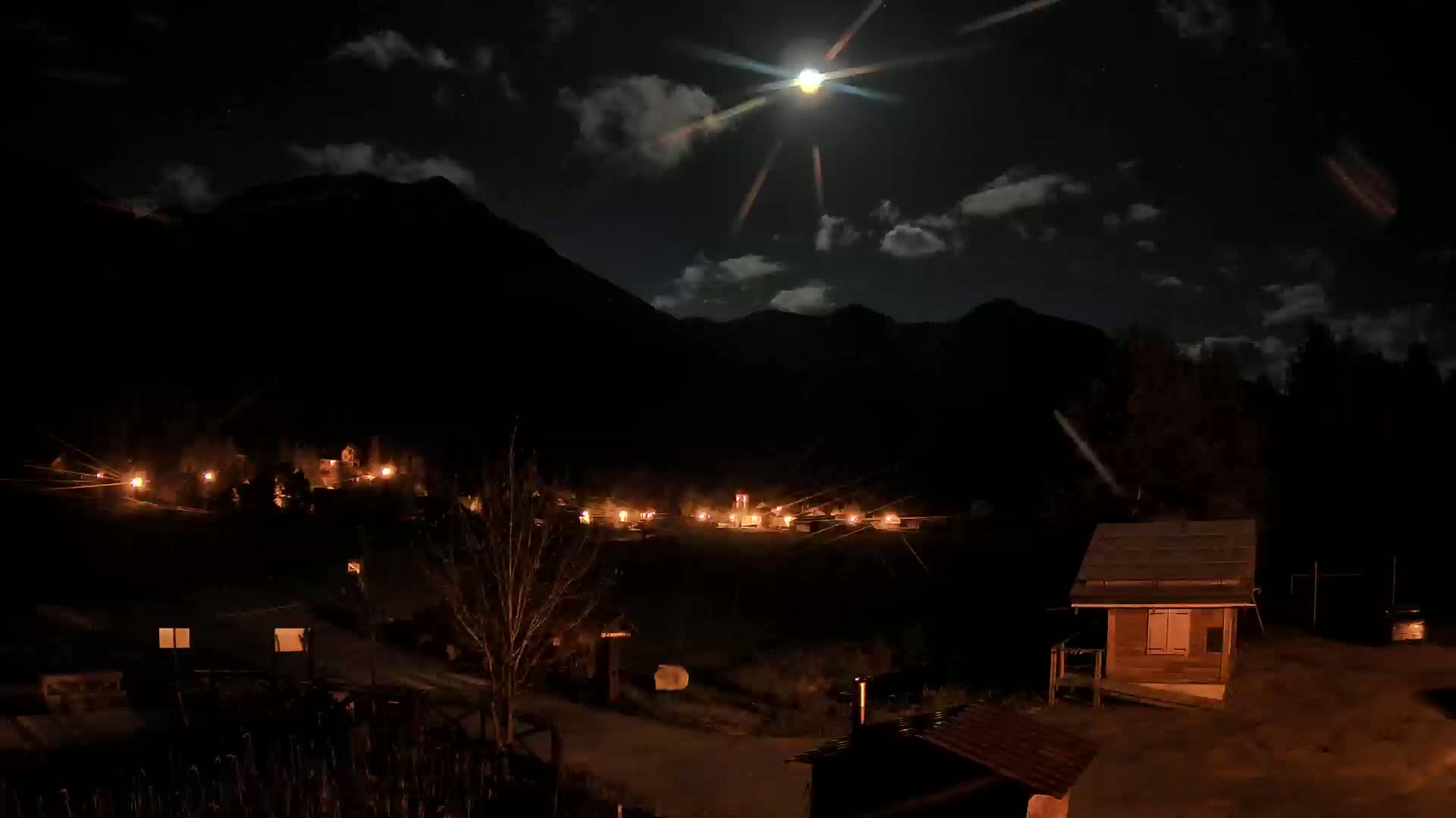 A moonlit night scene shows a small village nestled in a valley between dark mountains, with lights illuminating buildings and a sparsely wooded area.