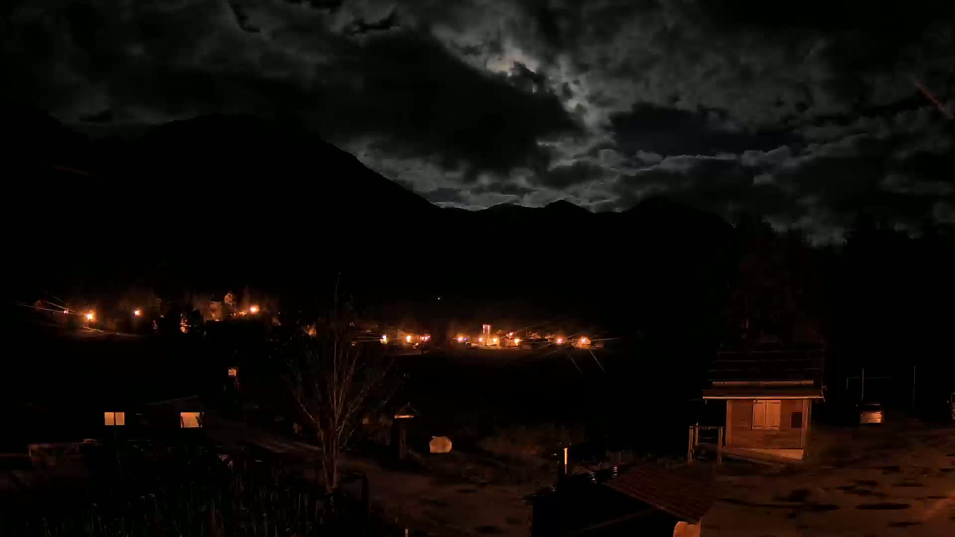 A moonlit night scene shows a small village nestled in a dark mountain valley under a cloudy sky.
