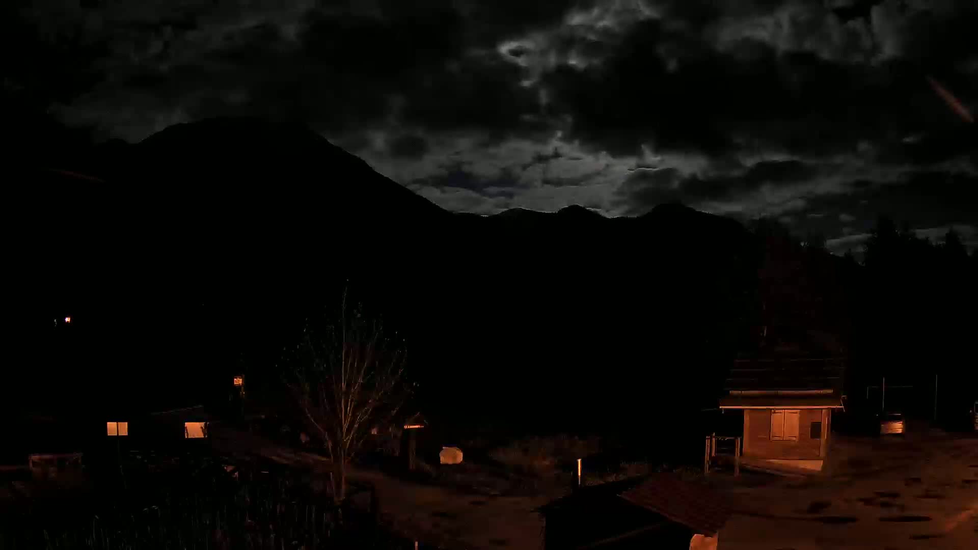 At night under a dark, cloudy sky, illuminated buildings are nestled at the base of a silhouetted mountain range.