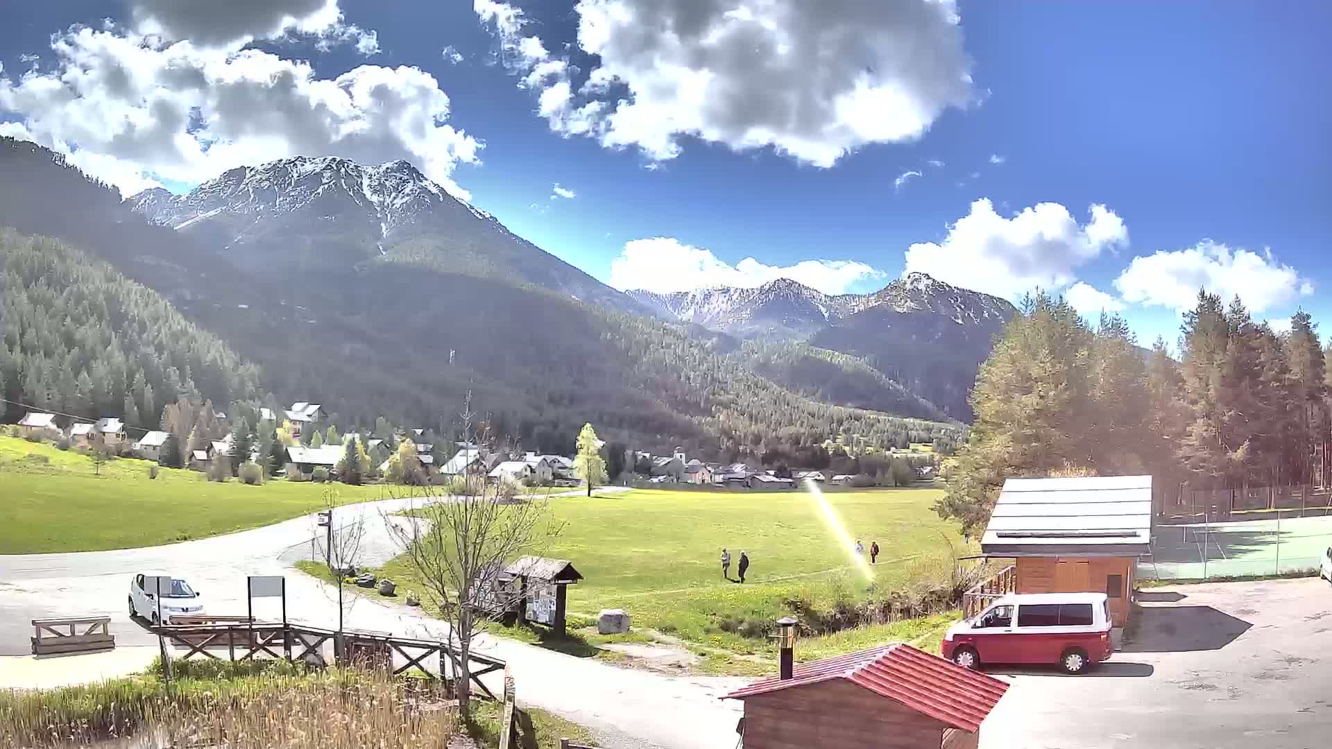 A sunny day with partly cloudy skies shows a small village nestled in a valley between snow-capped mountains, with a few people walking on a grassy field and several vehicles parked nearby.
