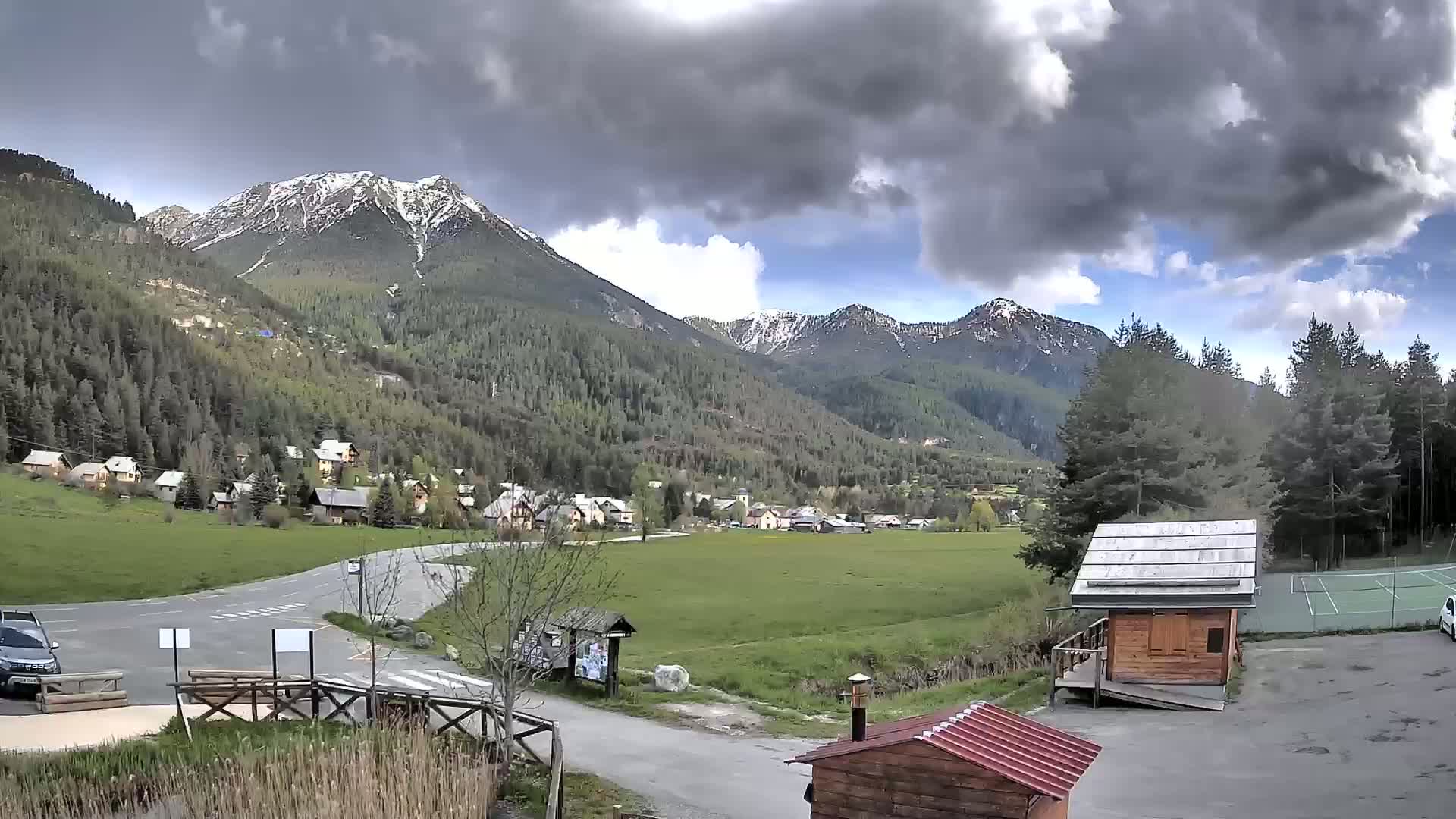 A small village nestled in a valley between snow-capped mountains under a partly cloudy sky.