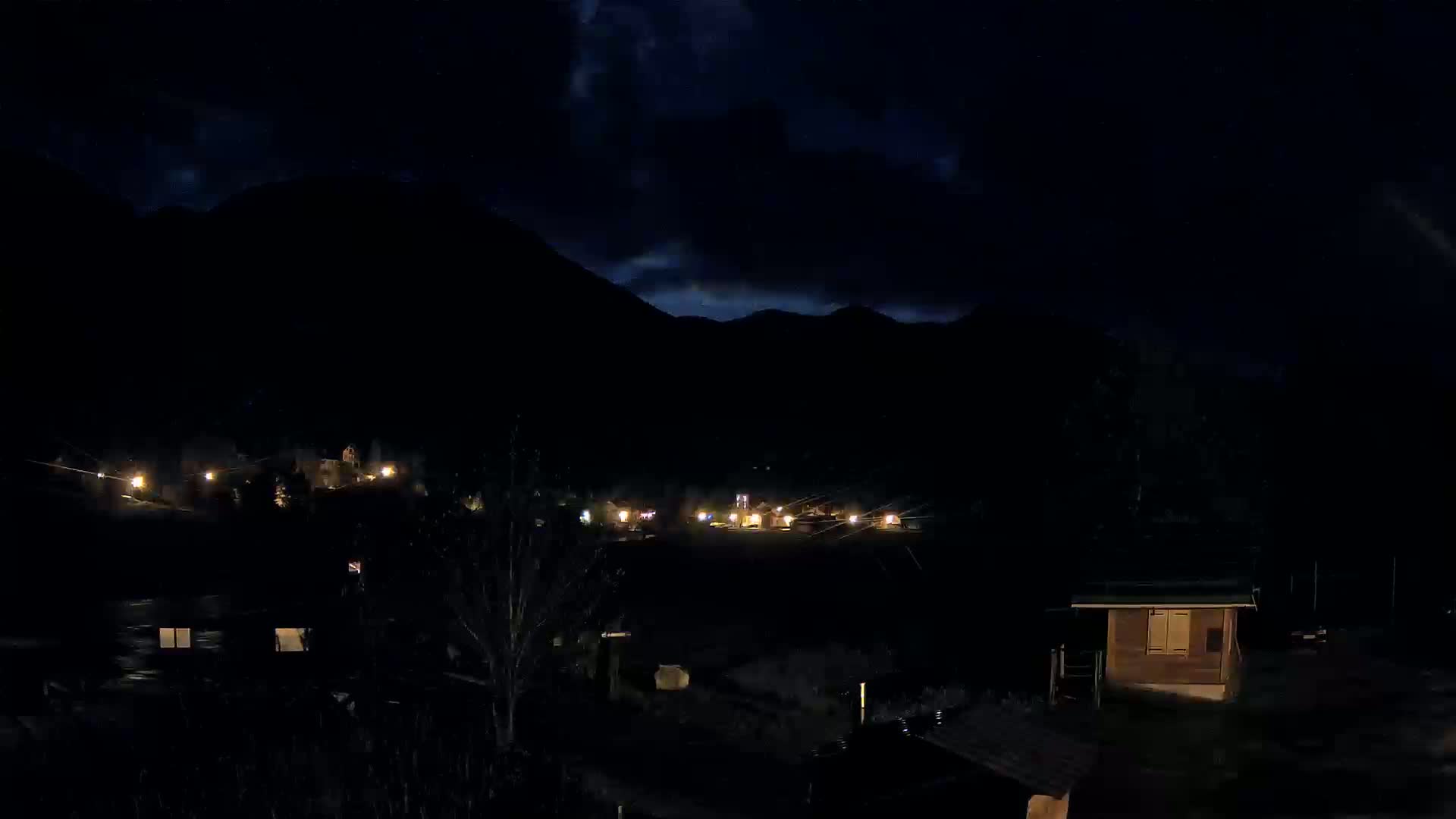 A nighttime view of a small town nestled in a valley between dark mountains under a cloudy sky.