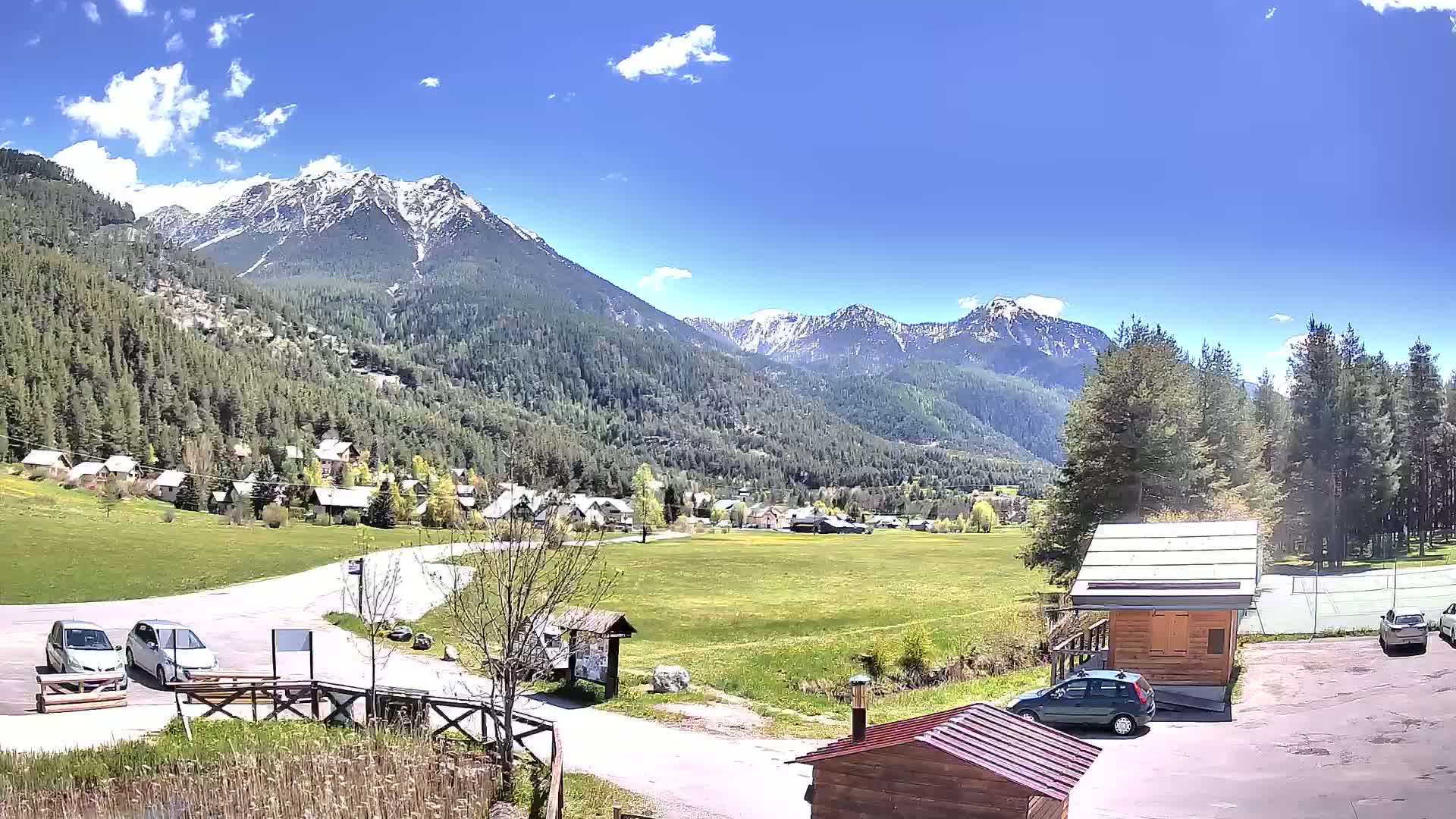A sunny day reveals a village nestled in a valley surrounded by snow-capped mountains and lush green fields.