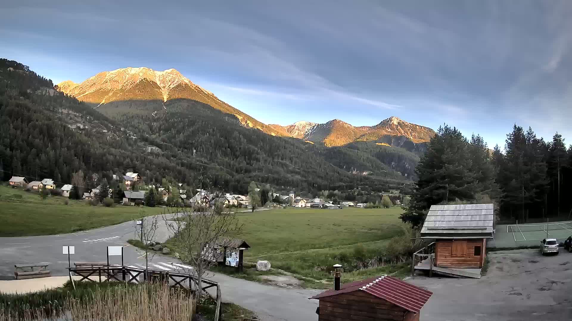 A small village nestled in a valley between snow-capped mountains under a partly cloudy sky.