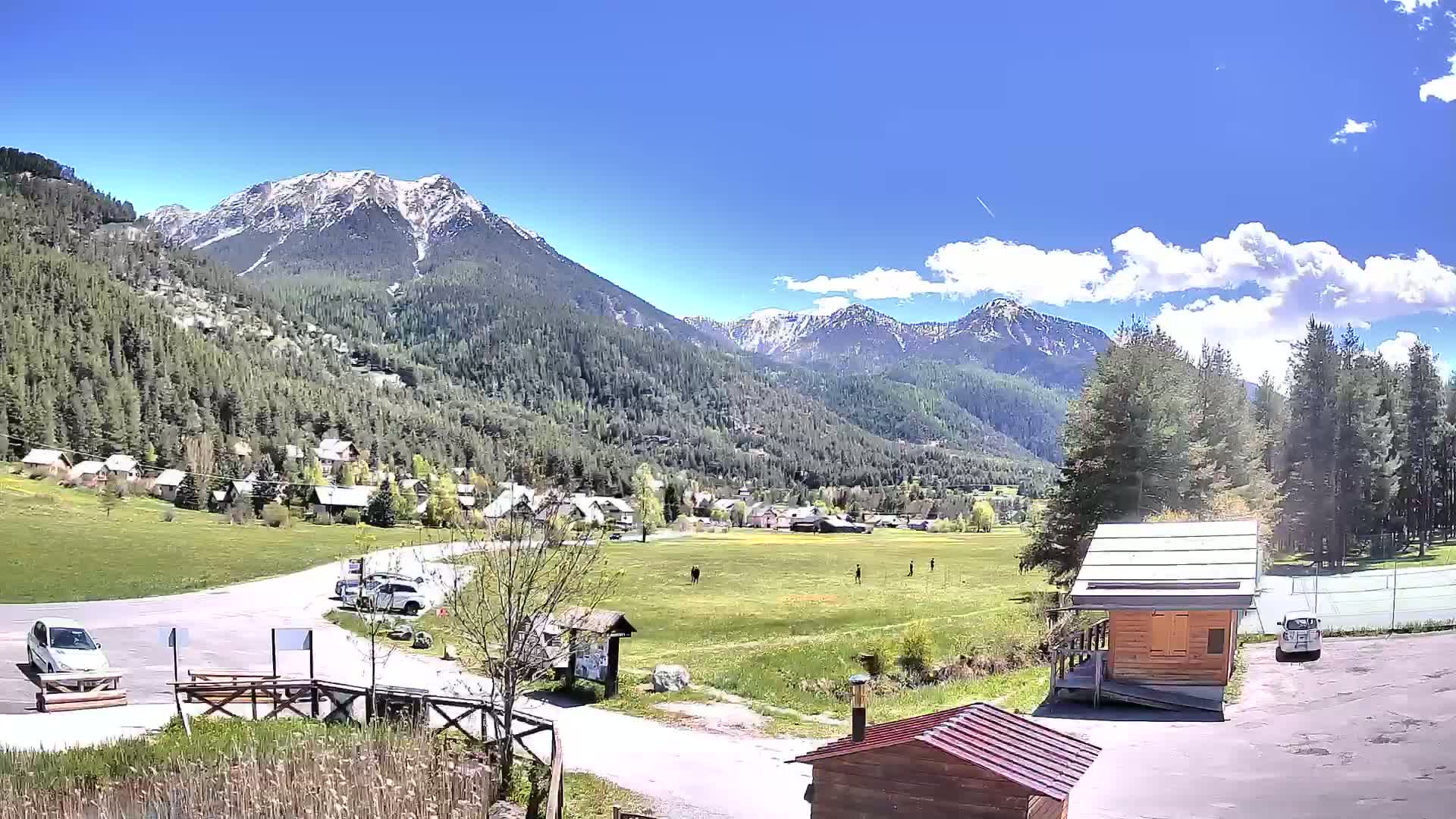 A sunny day in a mountainous valley shows a small village nestled among green fields and evergreen forests, with snow-capped peaks rising in the background.