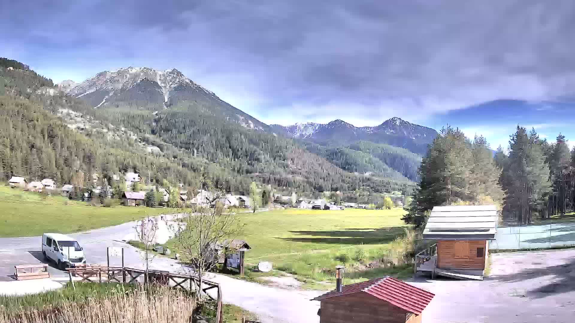A partly cloudy day reveals a valley town nestled amongst a range of snow-capped mountains, with a white van parked near small wooden structures.