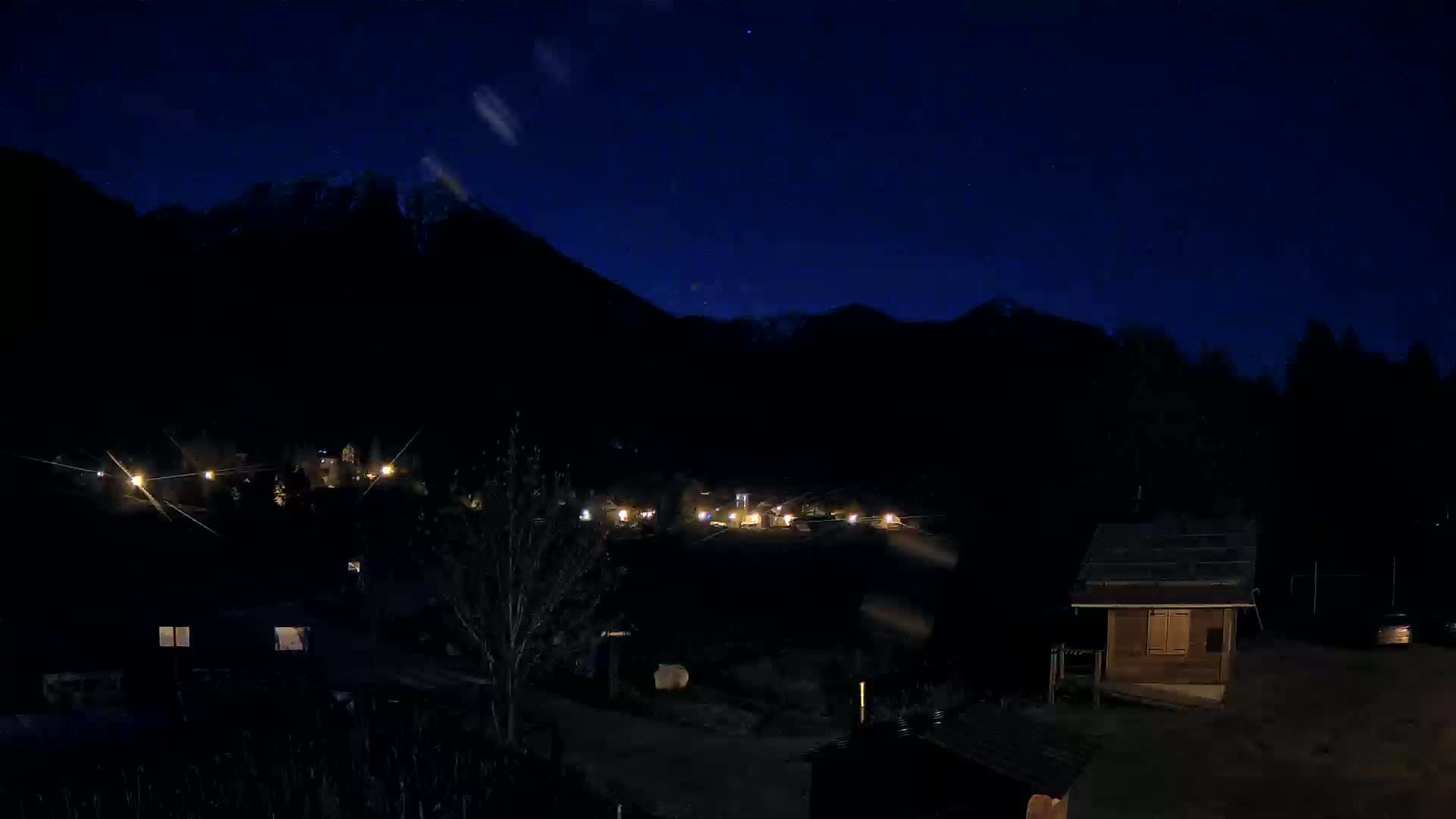 A nighttime view of a small village nestled in a valley, illuminated by scattered lights, with dark mountains rising in the background under a clear, starlit sky.