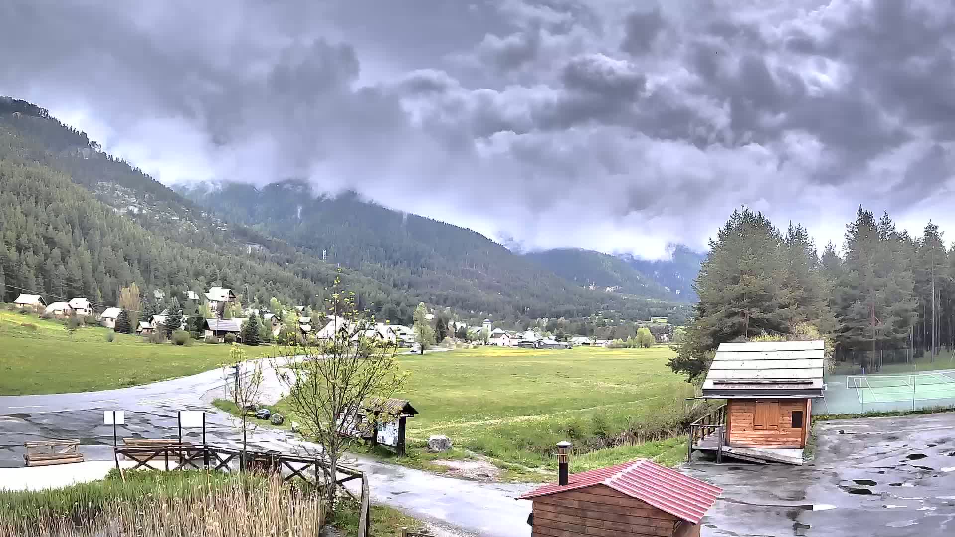 A small village nestled in a valley between mountains under a cloudy, possibly stormy sky, with a road leading to it and small wooden structures nearby.