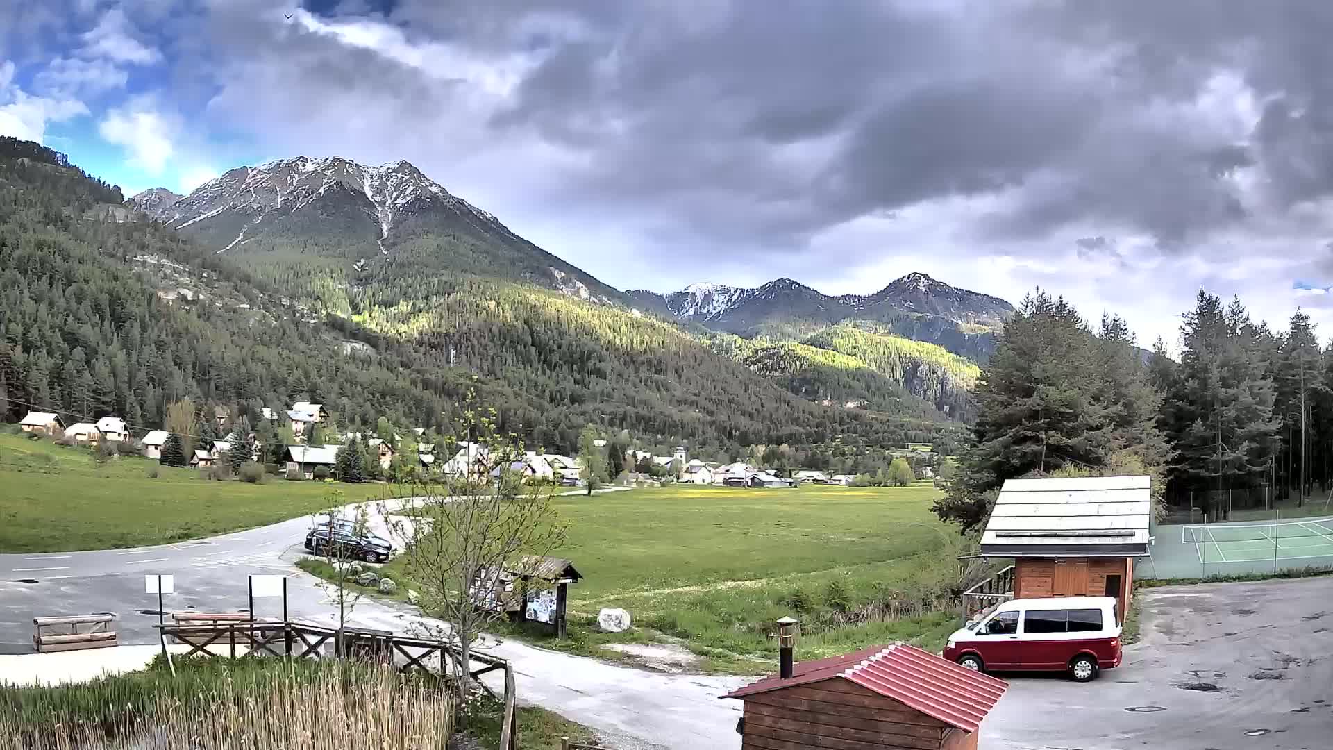 A small village nestled in a valley between snow-capped mountains under a partly cloudy sky, with a van parked near a tennis court.