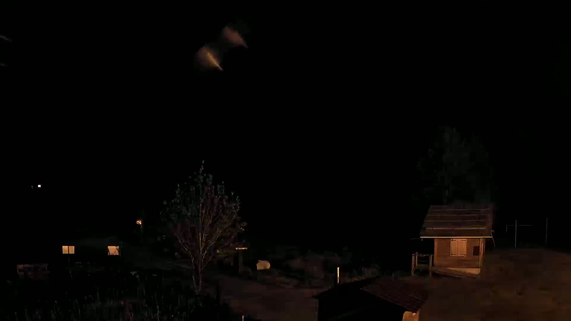 A nighttime scene shows a small wooden structure and a tree in a dark yard, with a faint light in the sky.