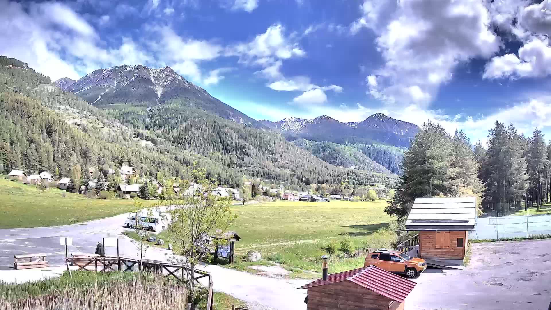 A partly sunny day shows a mountain valley with a small village nestled amongst green fields and forests, a road leading to a parking area with a few cars and small buildings nearby.