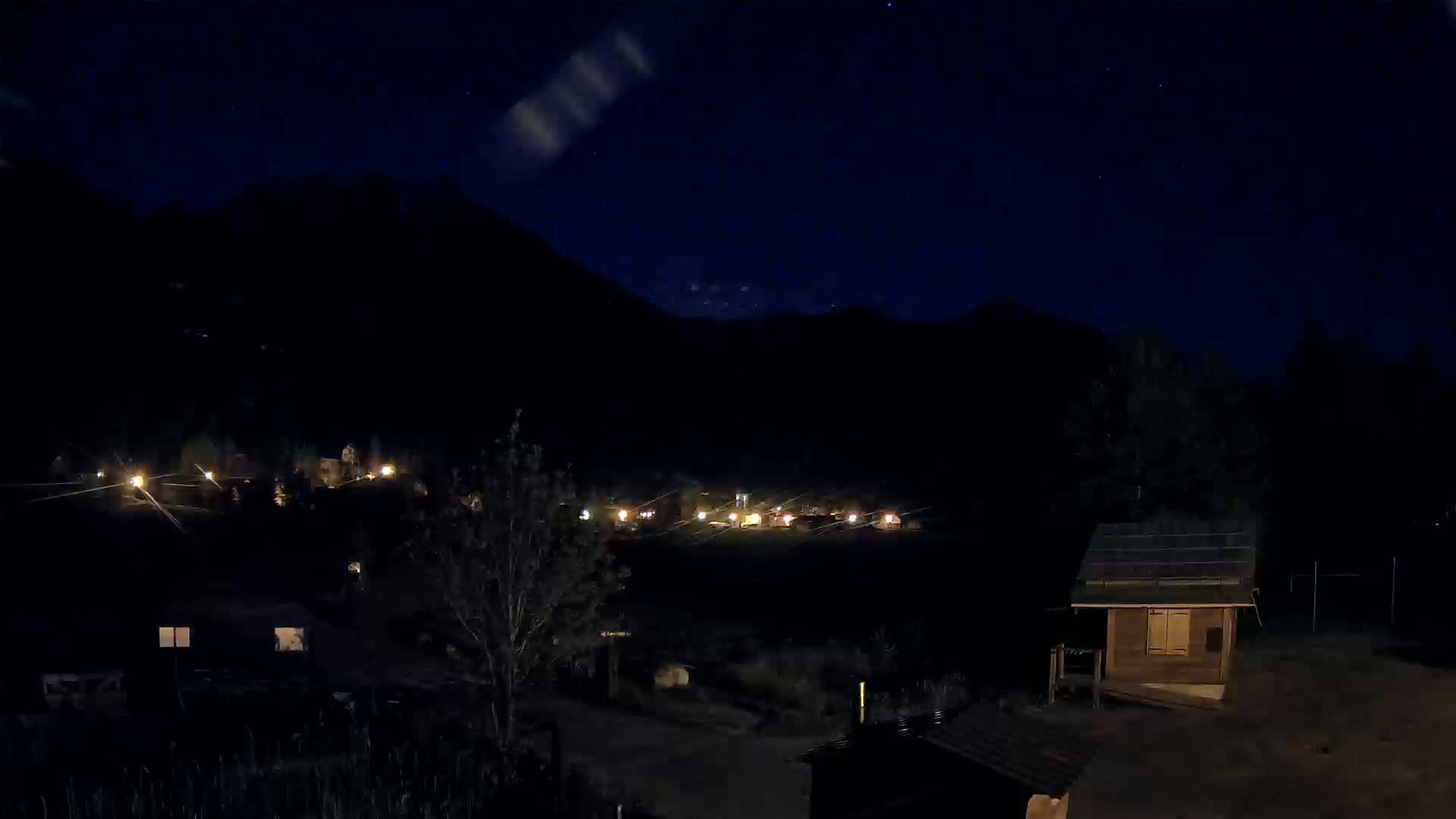 A nighttime view of a small settlement nestled in a valley between dark mountains under a clear, starry sky.
