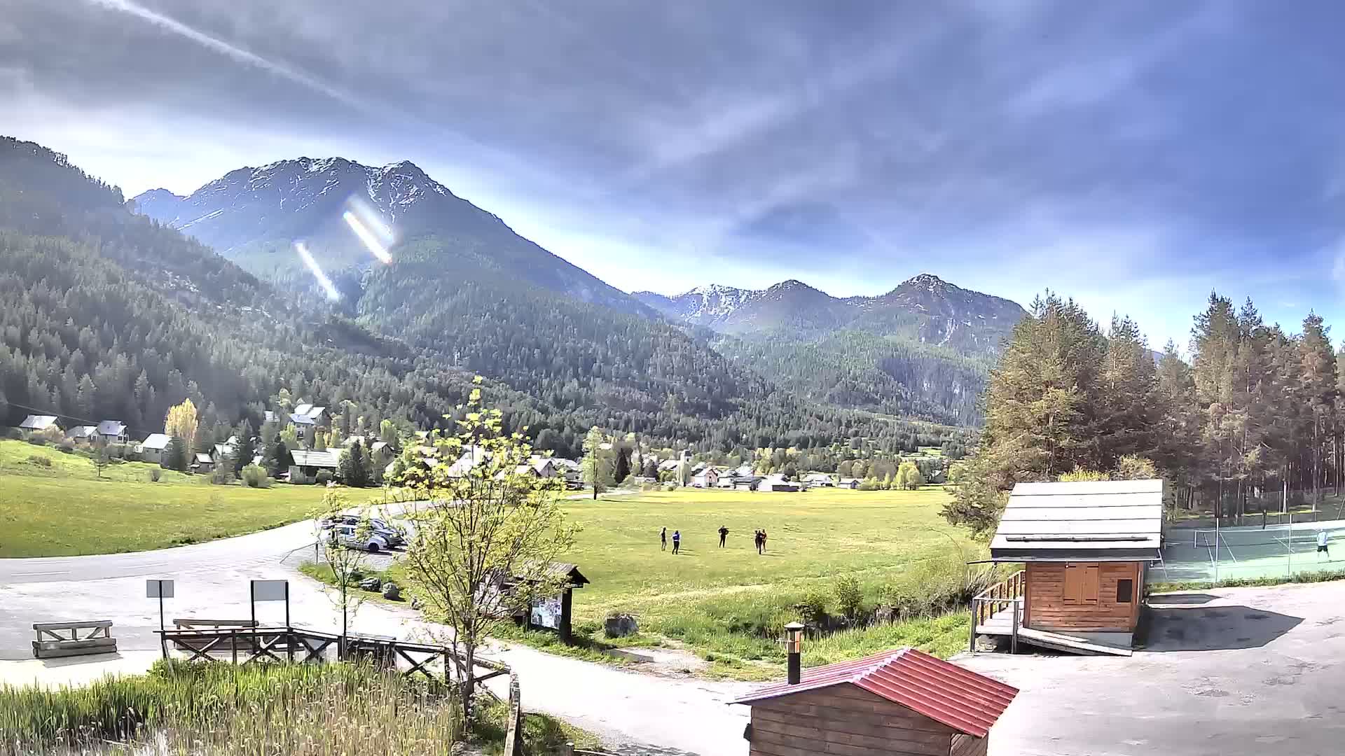 A sunny day reveals a small village nestled in a valley between snow-capped mountains, with a few people standing in a field and a small wooden structure in the foreground.