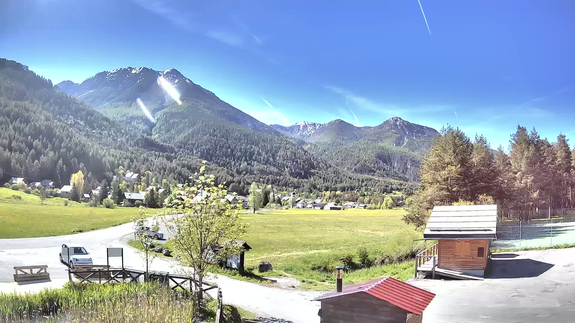 A sunny mountain valley scene shows a small village nestled among green fields and coniferous forests, with snow-capped peaks in the background.