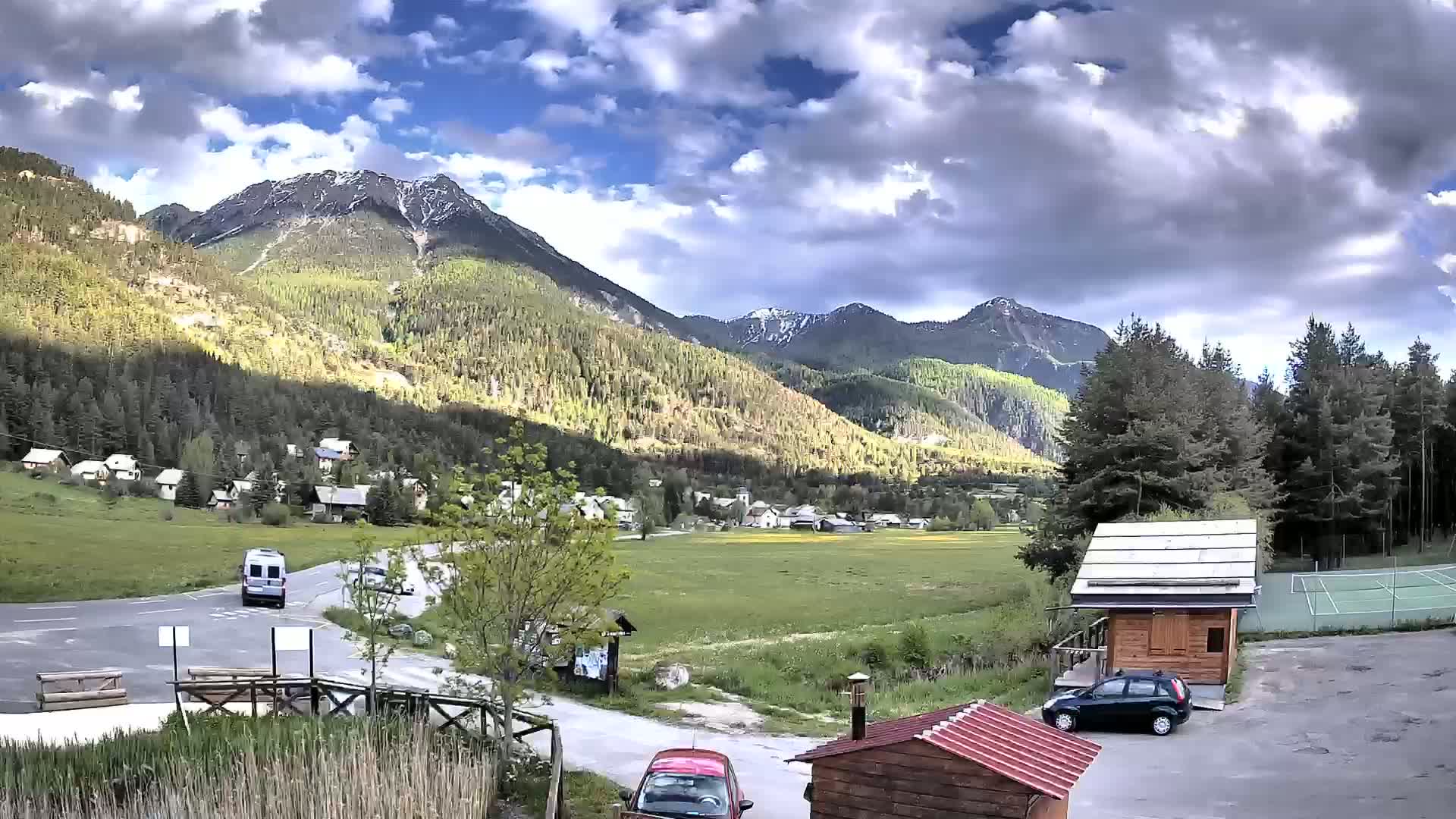 A partly cloudy day shows a small village nestled in a valley between snow-capped mountains, with a few cars parked near small wooden buildings and a tennis court.