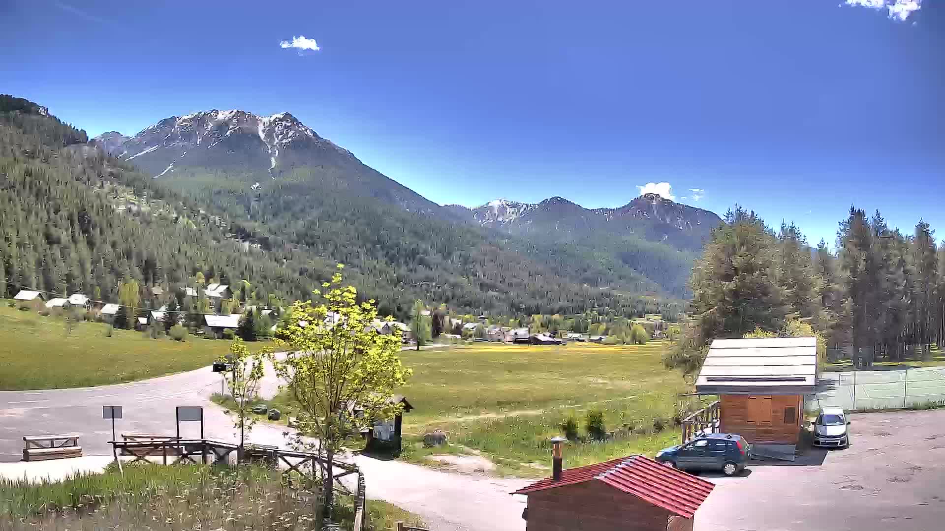 A sunny day reveals a mountain valley village nestled amongst snow-capped mountains and green forests, with a parking area containing several cars near a small wooden building.