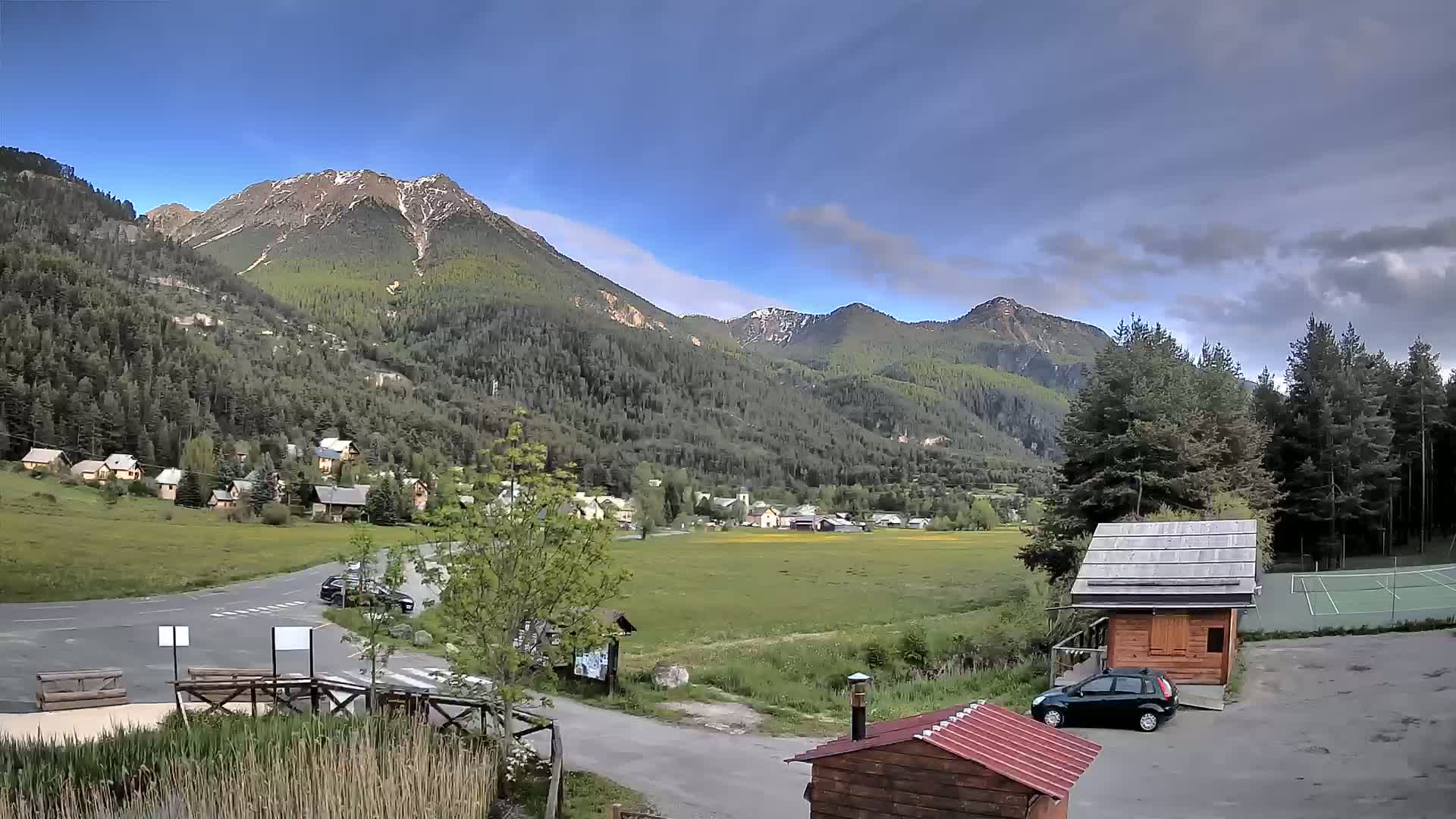 A mountain village nestled in a valley under a partly cloudy sky, with a small parking area and a tennis court visible in the foreground.