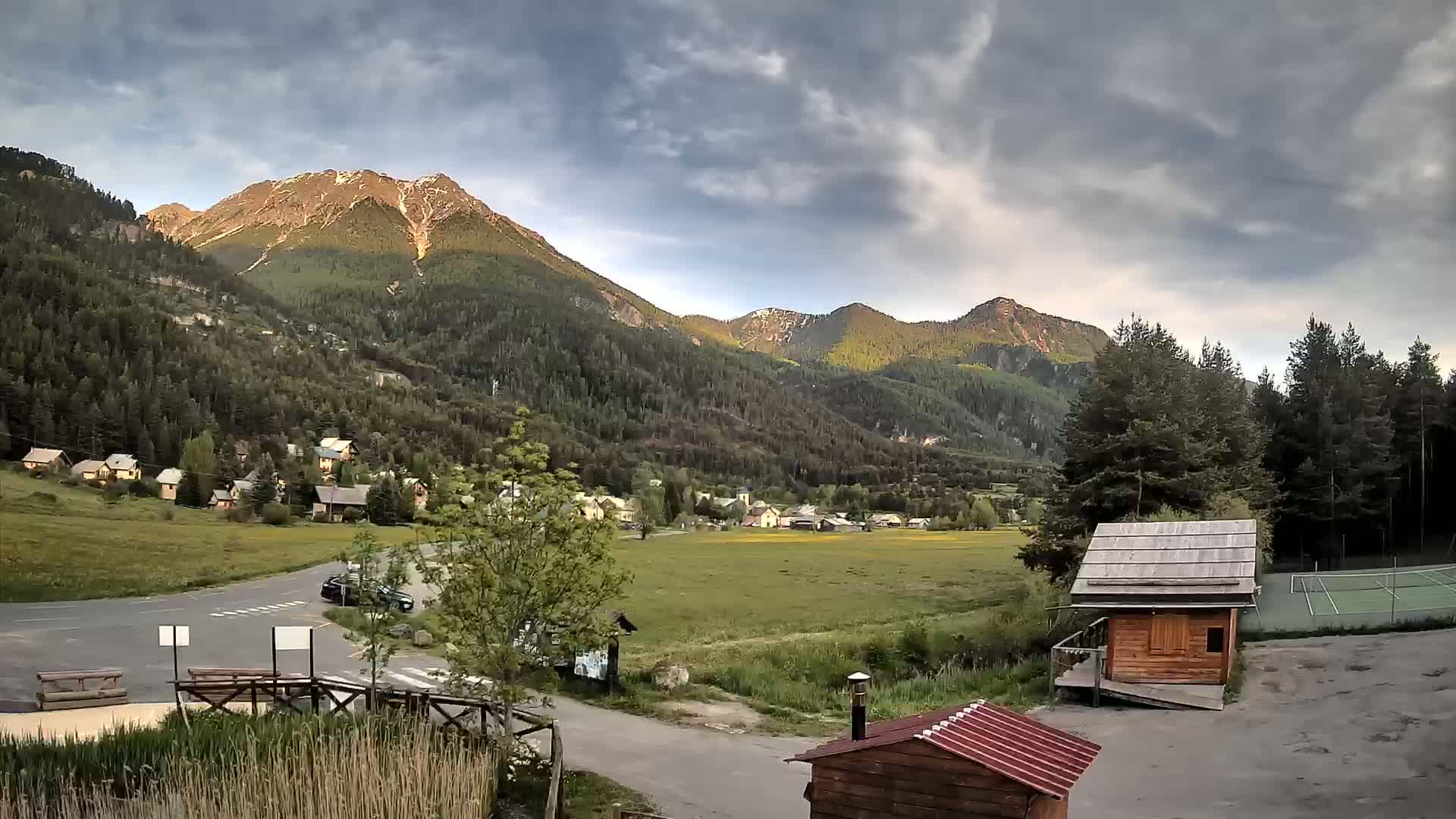 A mountain village nestled in a valley under a partly cloudy sky, with a tennis court and small wooden buildings nearby.