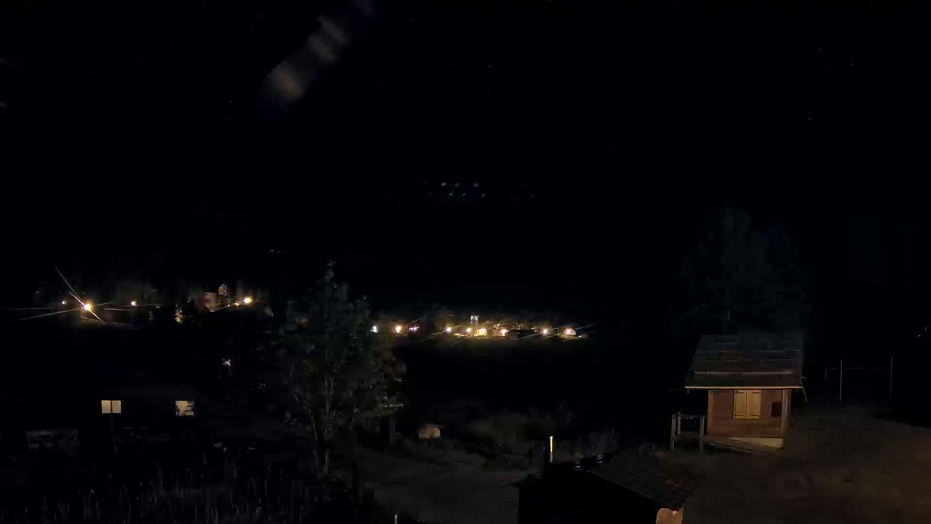 A nighttime view of a small settlement with scattered lights, a small wooden structure in the foreground, and a dark sky speckled with stars.