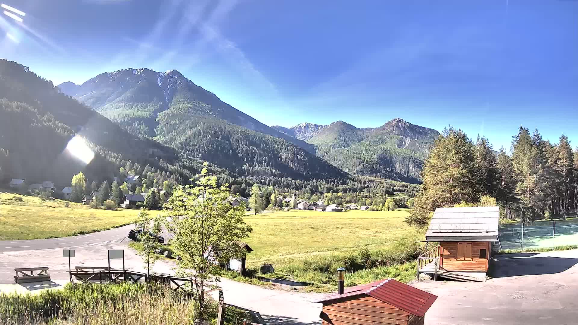 A sunny day reveals a valley nestled between snow-capped mountains, with a small village and a few structures visible in the foreground.