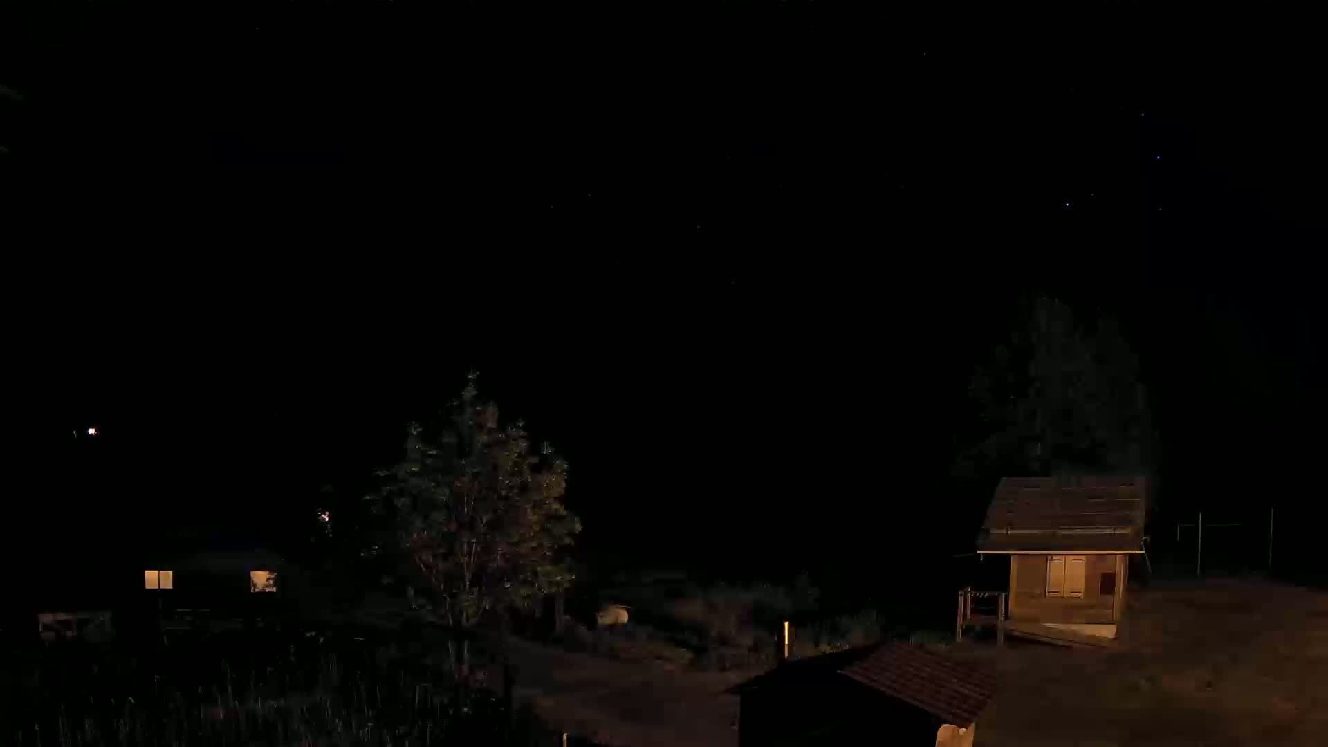 At night, under a clear, starry sky, two small buildings are illuminated, surrounded by trees and shrubs.