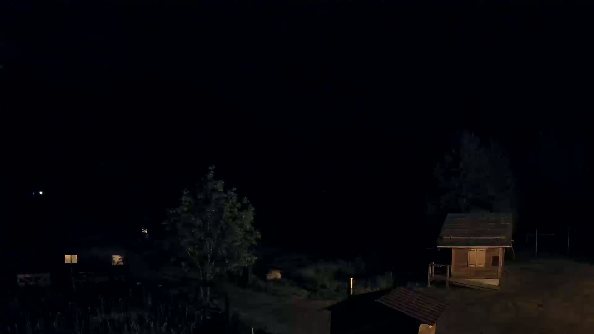 At night, a small wooden cabin and other structures are visible in a dark, sparsely lit outdoor area with trees.