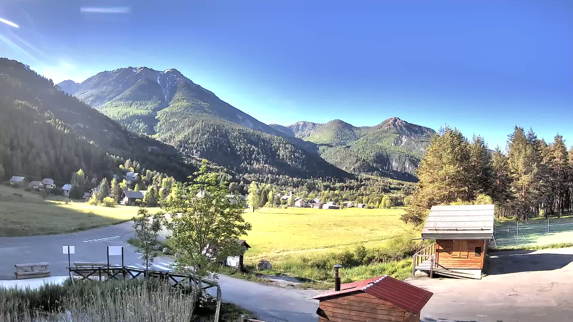 A sunny day reveals a mountain valley village nestled amongst green hills and coniferous trees, with two small wooden structures in the foreground.
