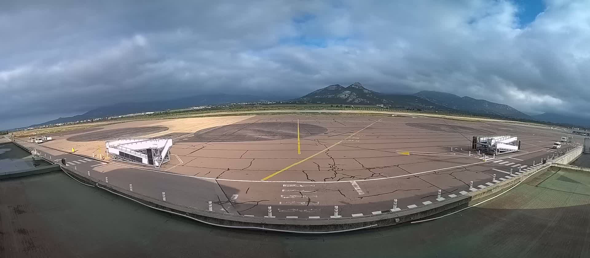 An airport tarmac with two jet bridges is visible under a cloudy sky, with mountains in the background.