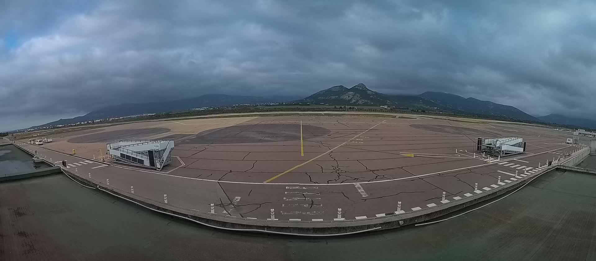 An empty airport tarmac under a cloudy sky, with mountains visible in the distance and two jet bridges positioned on either side of the apron.