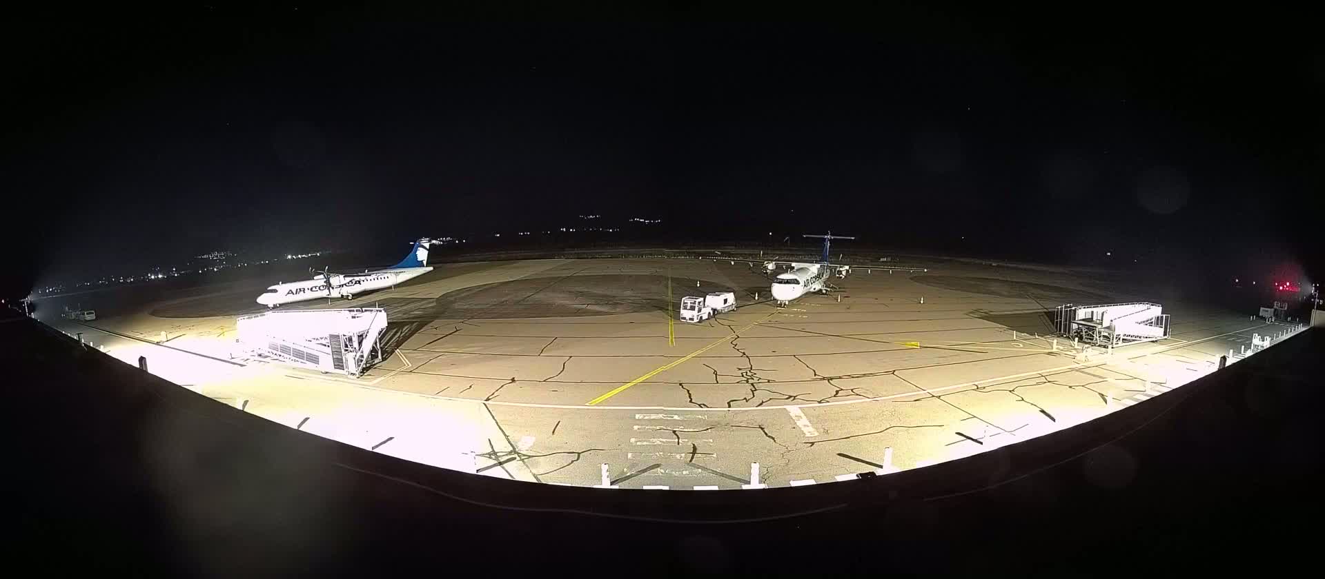 Two airplanes are parked on a tarmac at night, illuminated by lights, with ground support equipment nearby.