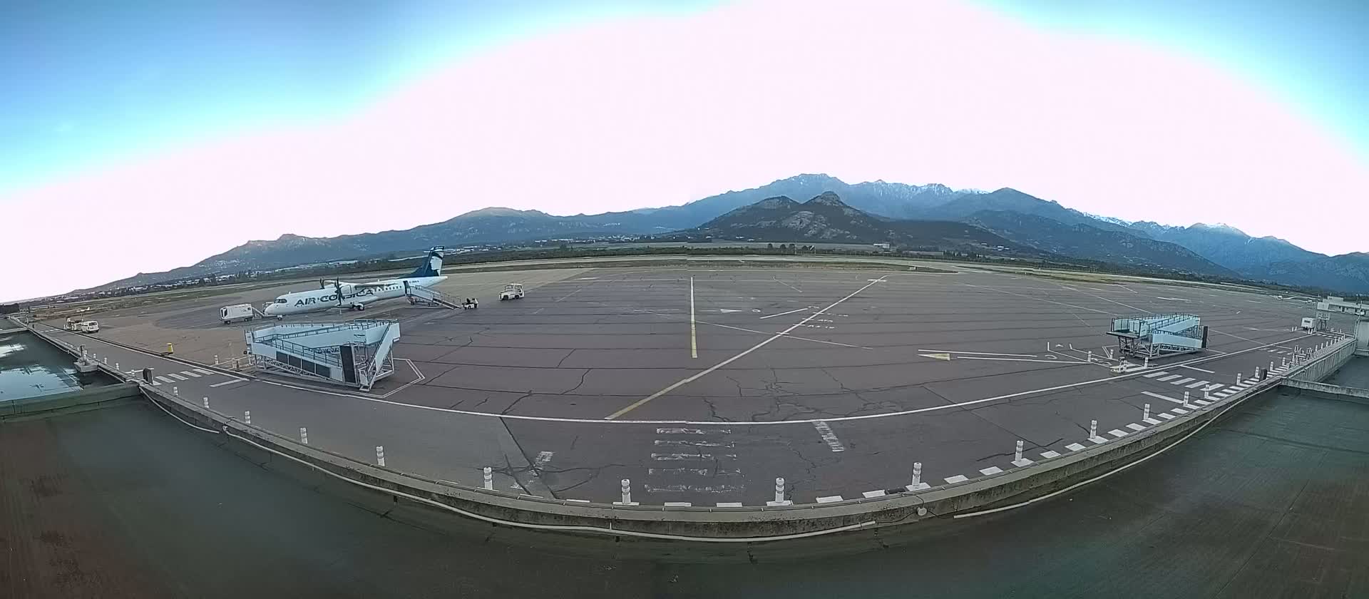 An airplane is parked at a gate on a tarmac, with a mountainous landscape visible in the background under a clear sky.
